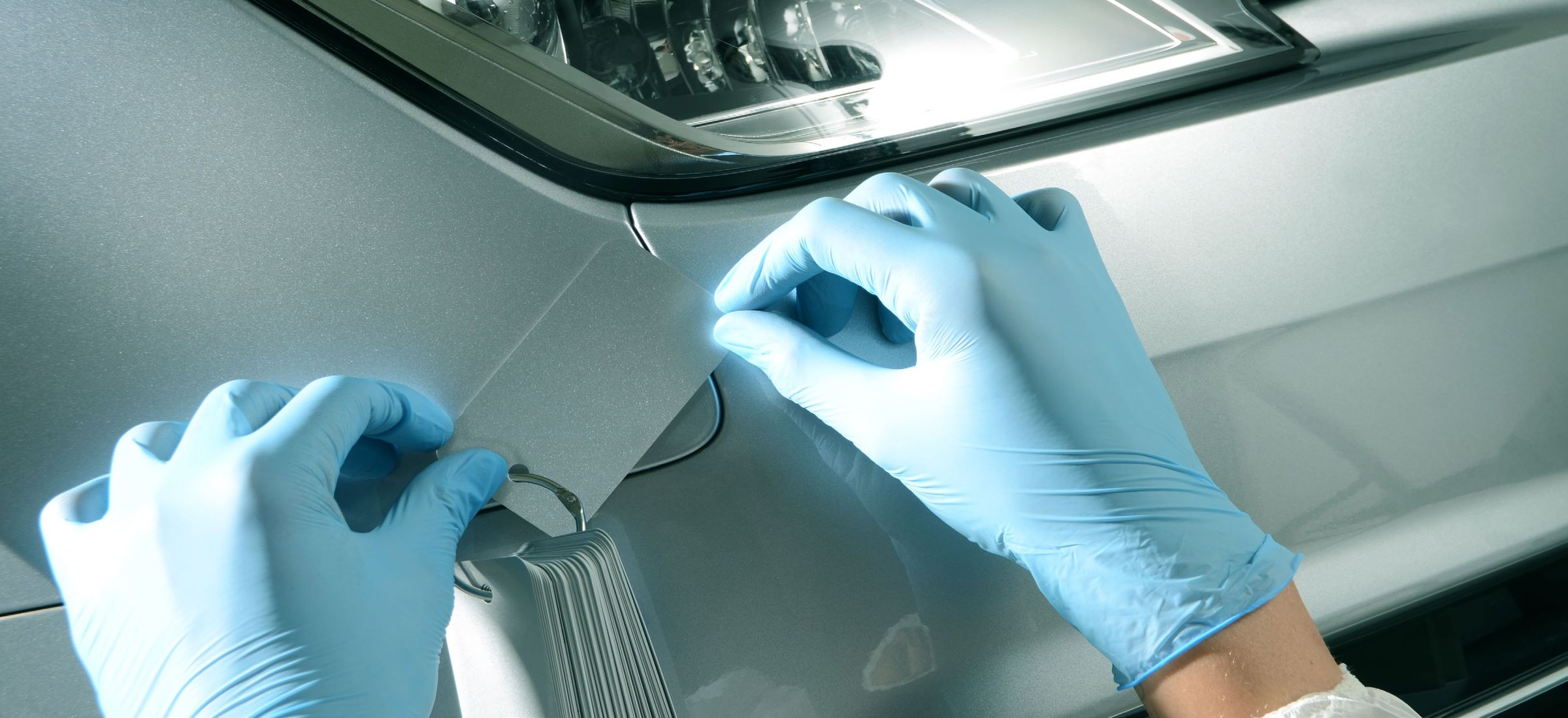 A gloved hand holding a color swatch against a silver car panel